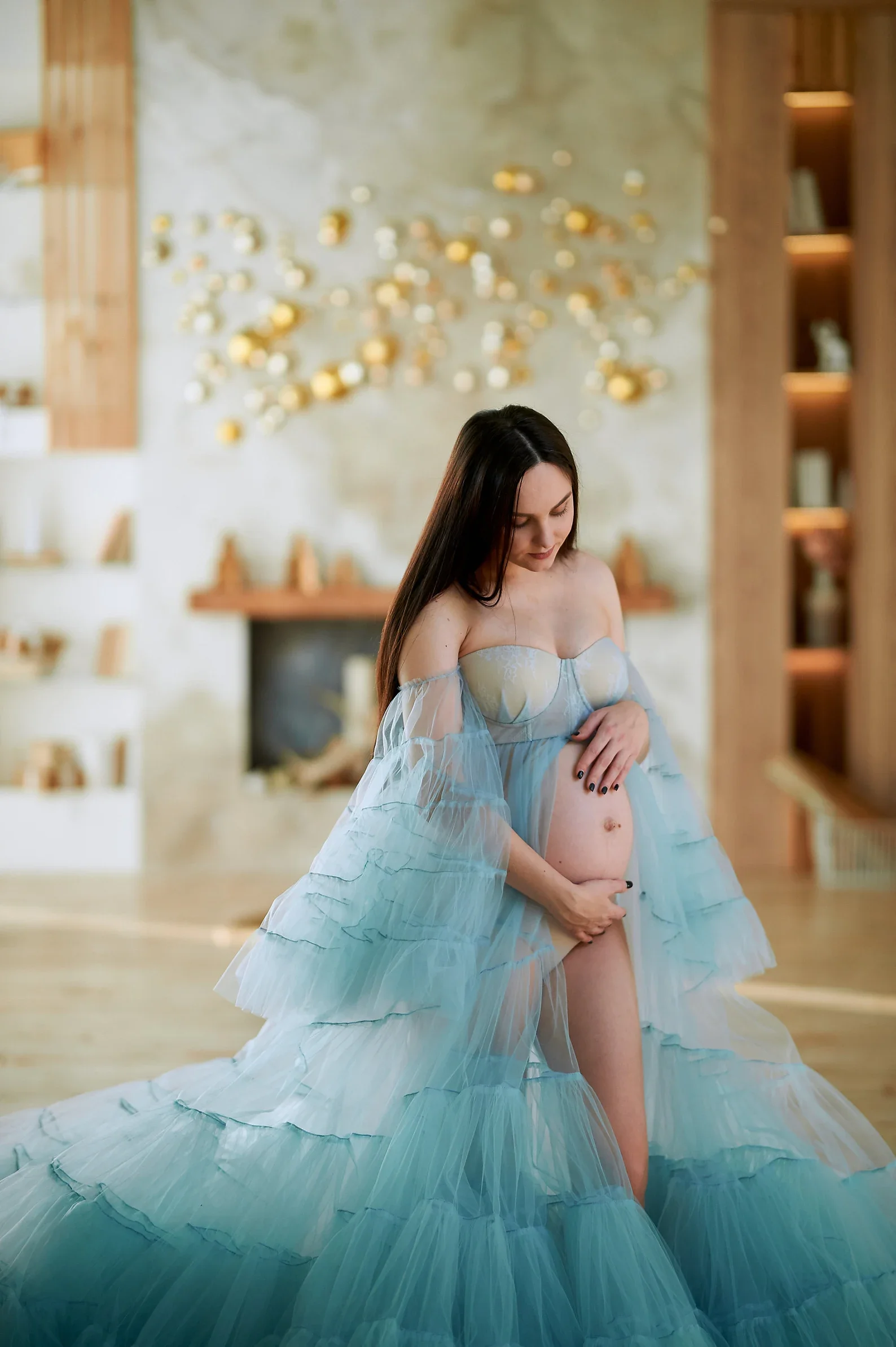 Pregnant woman in a flowy, sheer blue dress, gently holding her belly, standing in a warmly lit room.