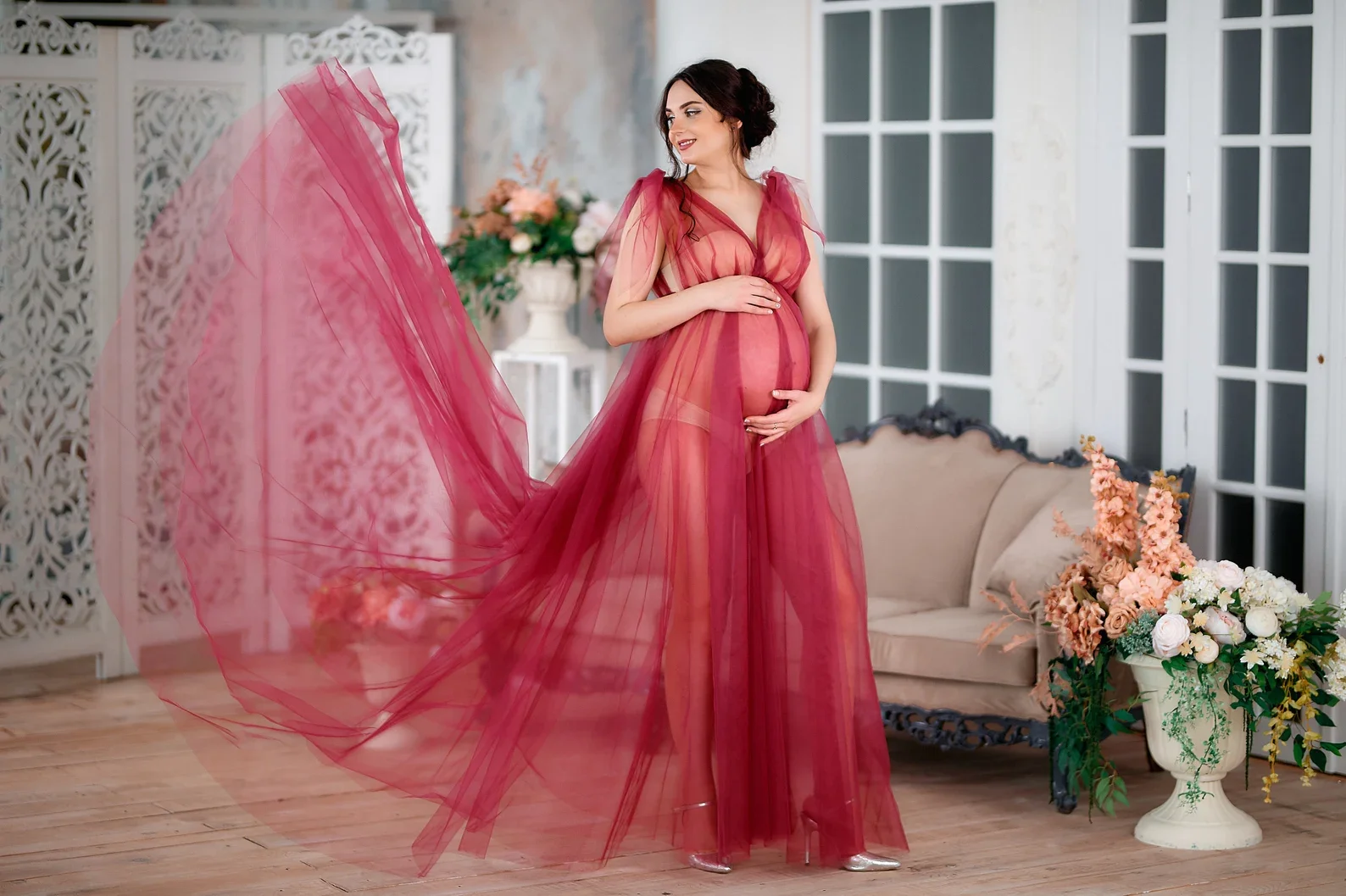 A pregnant woman in a sheer pink and red gown is posing indoors. She stands on a wooden floor near a beige sofa and floral arrangements in vases. The room has large windows and white decorative lattice panels.