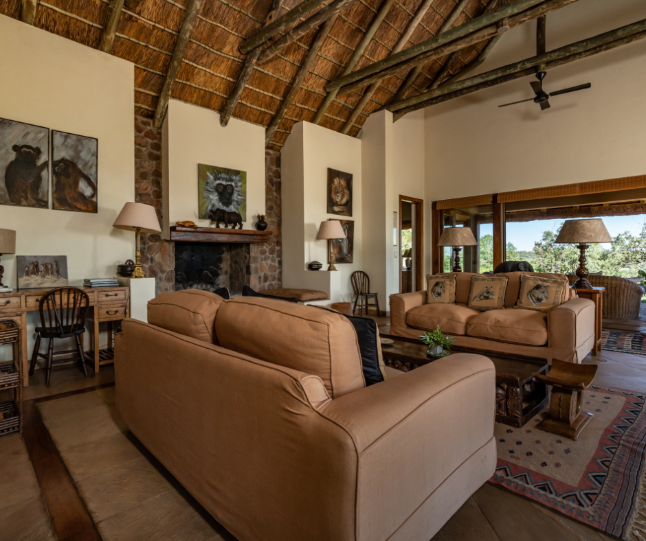 Living room with high thatched ceiling, stone fireplace, and large windows showing outdoor trees. Furnished with beige sofas, wooden coffee table, lamps, paintings of animals, and a ceiling fan.