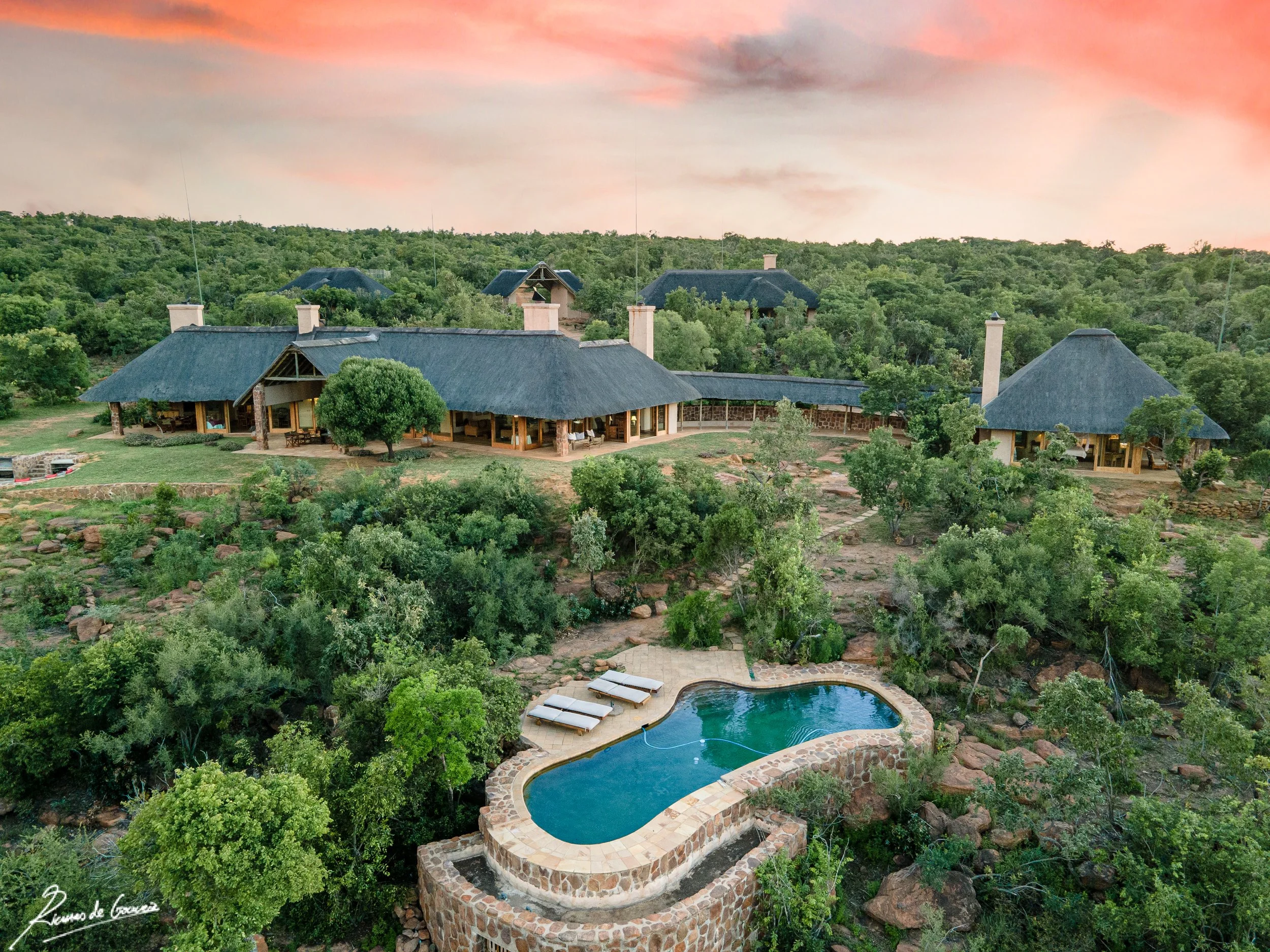A large rustic house with thatched roofs surrounded by trees, a stone-lined swimming pool with lounge chairs nearby, set in a lush green landscape at sunset.