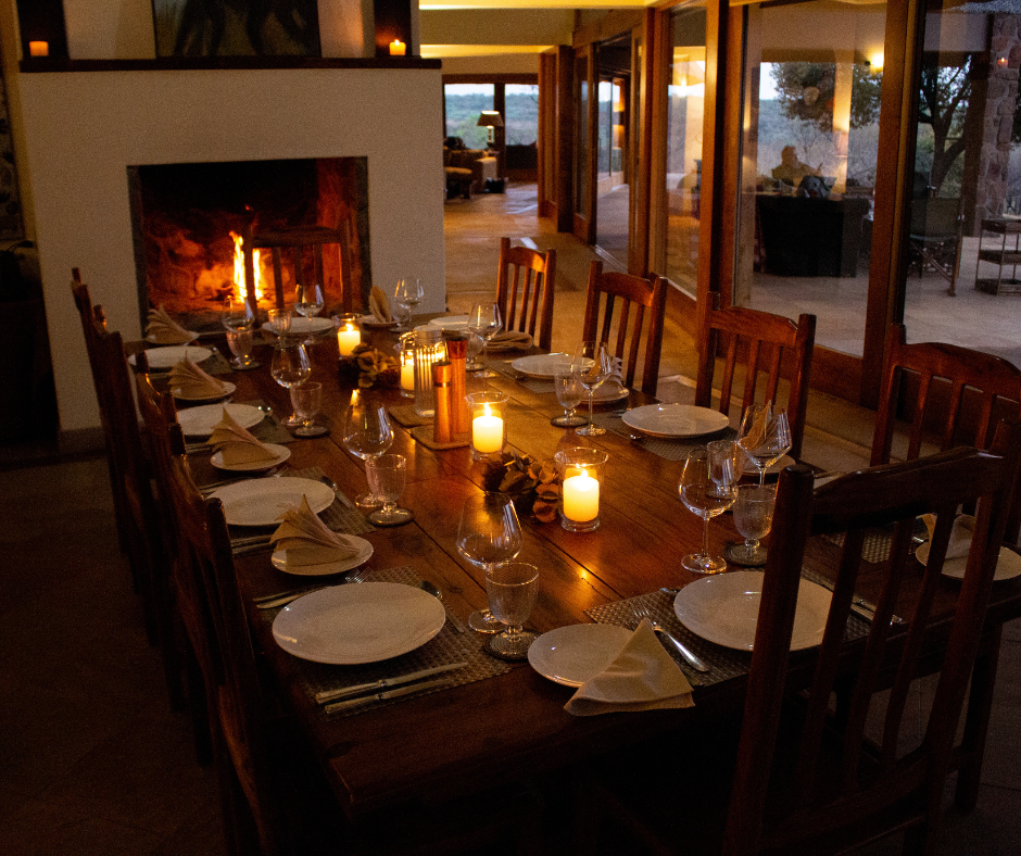 A dining room with a long wooden table set with plates, glasses, and napkins, lit by candles, with a fireplace in the background and large glass windows showing an outdoor patio and trees.