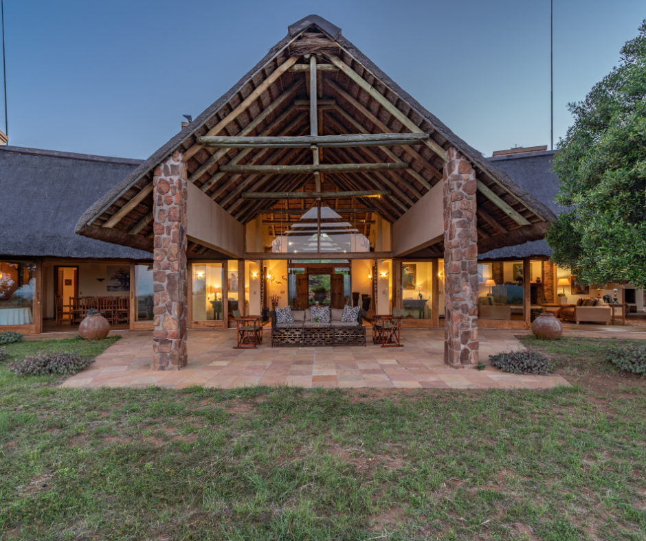 Front view of a house with a large open porch, stone pillars, and a thatched roof, illuminated from inside, with a grassy yard in the foreground and trees on the sides.