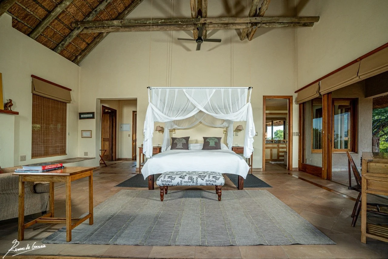 A spacious bedroom with a high, thatched ceiling and exposed wooden beams. It features a large bed with white linens and decorative pillows, a canopy frame, and a bench at the foot of the bed. There are wooden side tables with lamps, large windows wi