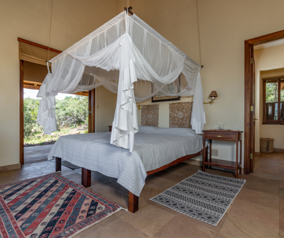 A bedroom with a four-poster bed, white canopy, patterned headboard, and a white bedspread. There are two rugs on the wooden floor, one colorful and one black and white. Two side tables and a window showing outdoor greenery are visible.
