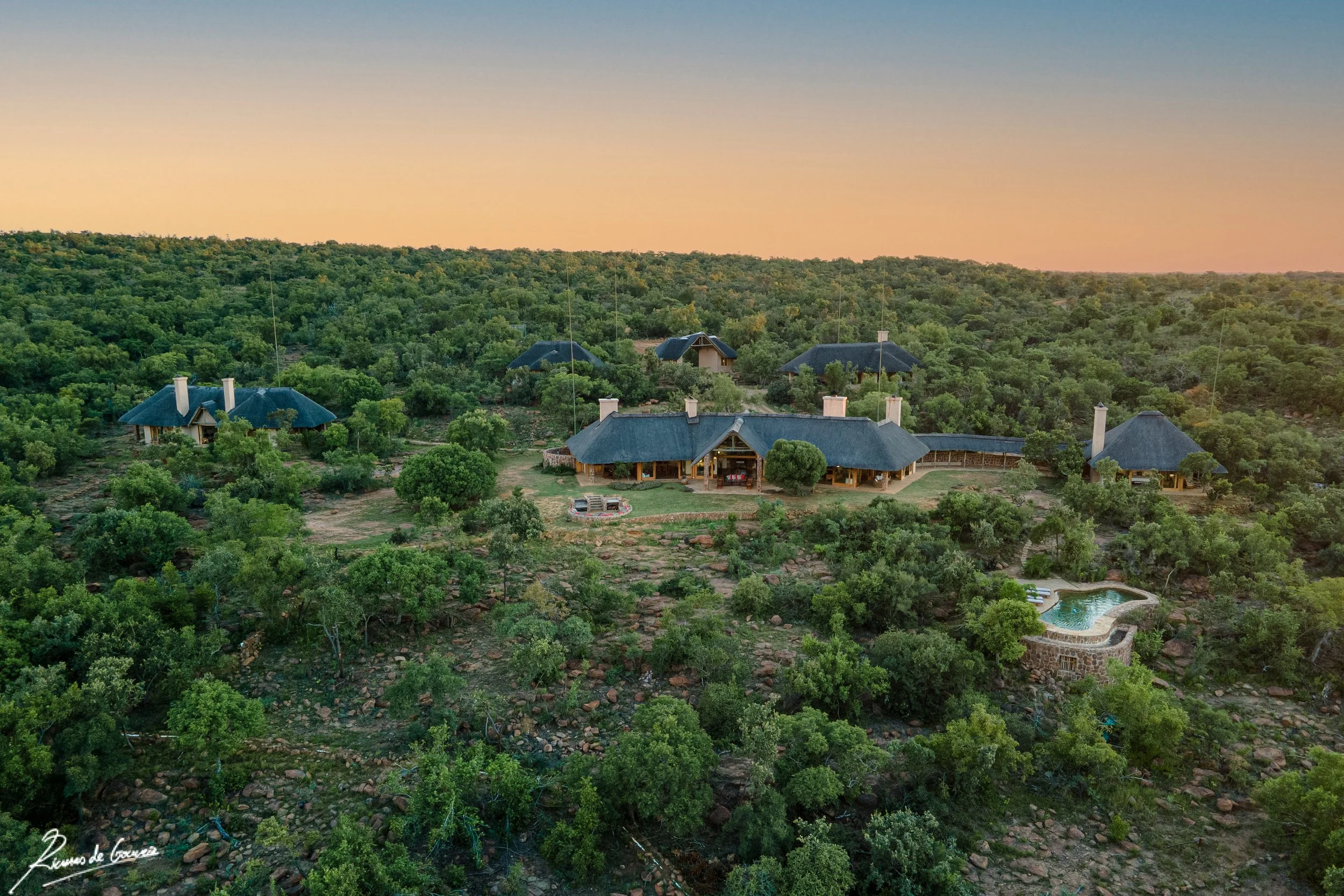 A large house and several smaller buildings are surrounded by lush green trees and shrubs on a hillside, with a sunset sky in the background. There is a swimming pool on the right side of the property.