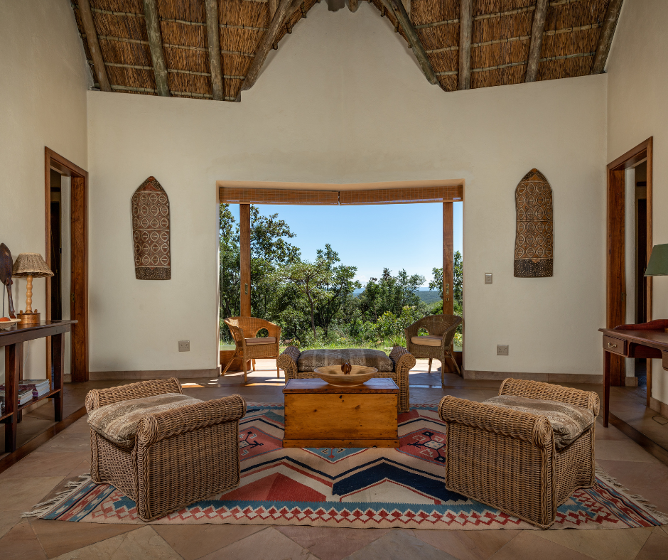 Living room with wicker chairs, wooden coffee table, colorful geometric rug, and view of trees and sky through a bay window.