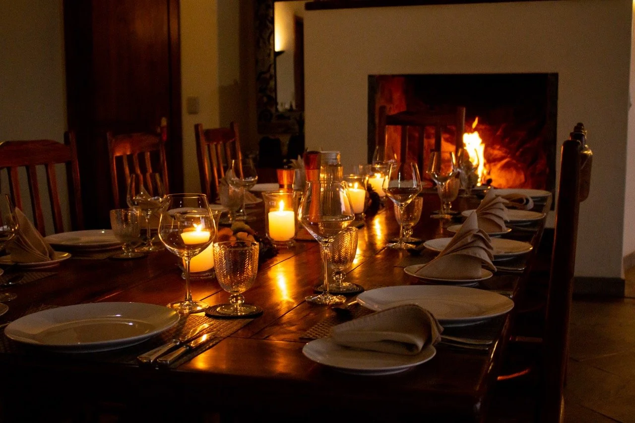 A dining table set for a meal with plates, napkins, glasses, and candles, in front of a lit fireplace.