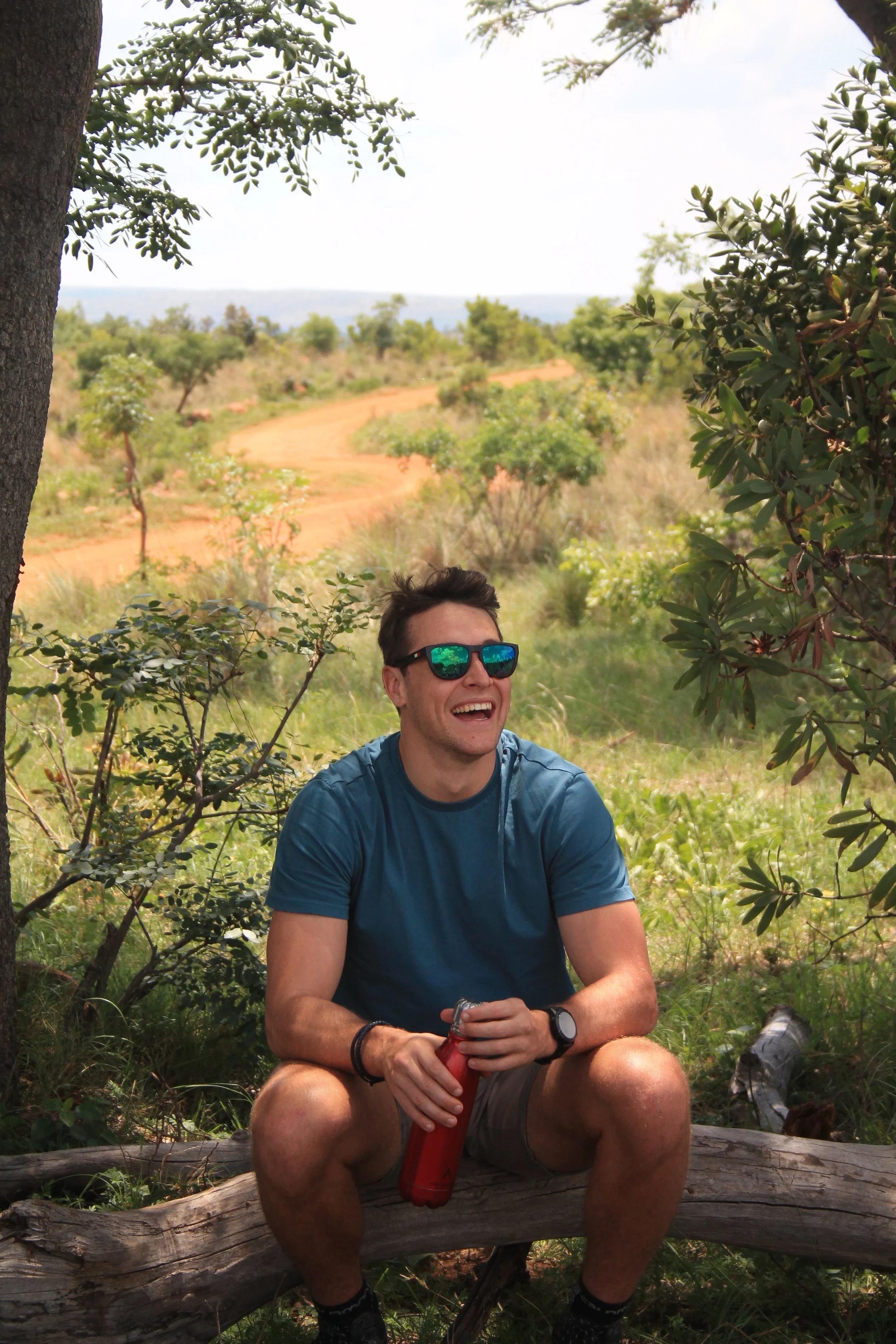 A young man sitting on a fallen log under a tree, laughing with sunglasses, holding a red water bottle, in a lush green outdoor setting with a dirt trail and open landscape in the background.