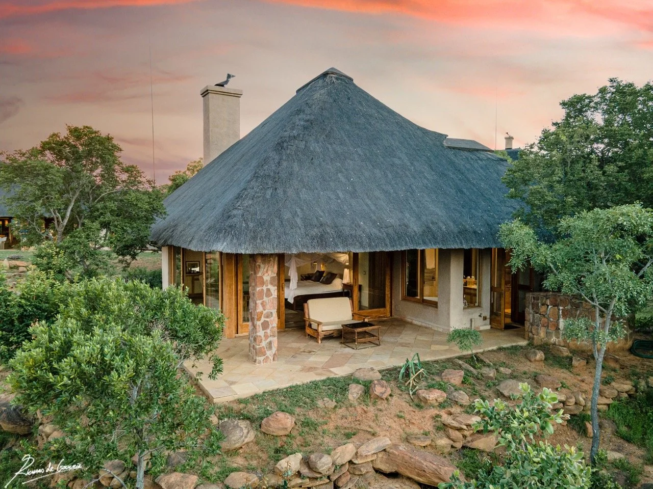 A rustic house with a thatched roof surrounded by greenery and rocks, sunset sky in background, interior lit up, with a sitting area visible through open doors.
