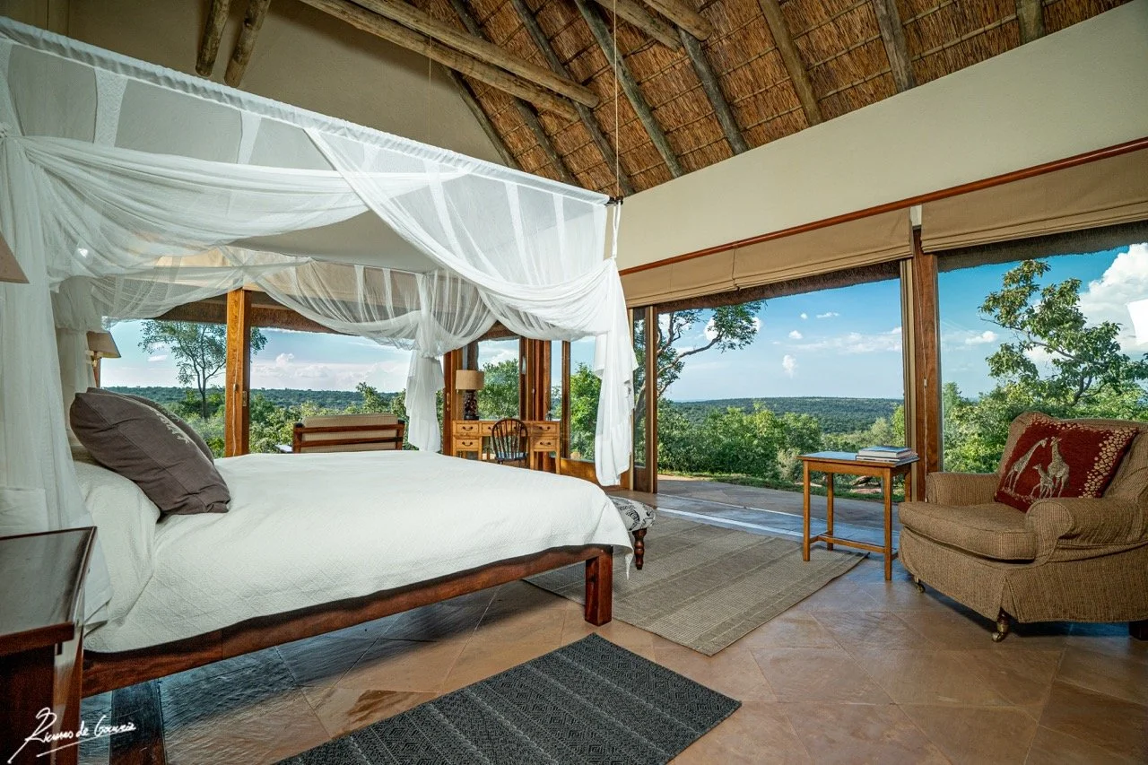 A bedroom with a large bed featuring a white quilt and dark pillows, a thatched roof, and large windows with a view of trees and blue sky. There is a sofa with a decorative pillow, a small side table, a desk with a chair, and a bench at the foot of the bed.