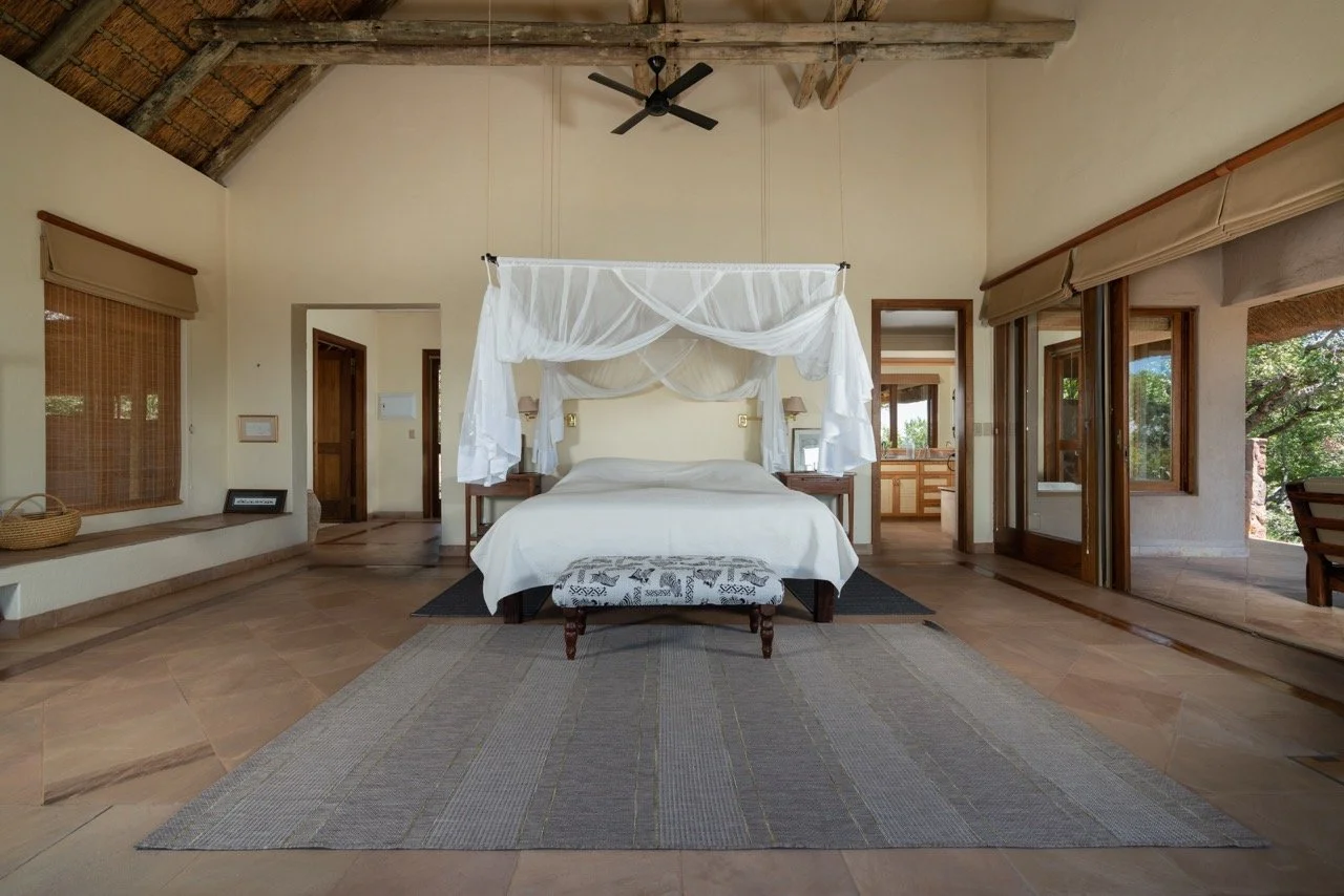 A spacious bedroom with high wooden ceilings and large windows, featuring a white bed with a canopy, a small bench at the foot, and a view of greenery outside.