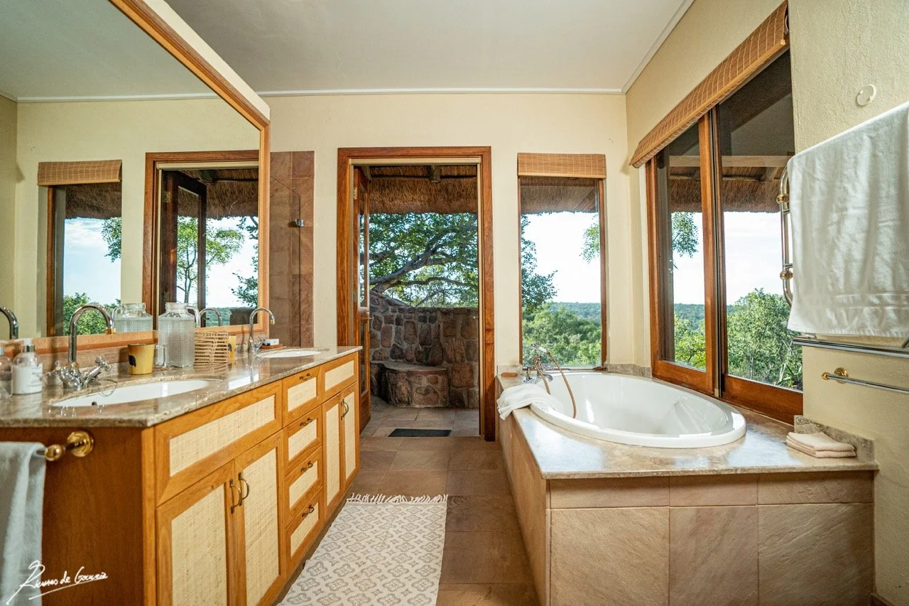 Bathroom with double sink vanity, large mirror, walk-in shower, and a bathtub next to large windows showing trees and greenery.