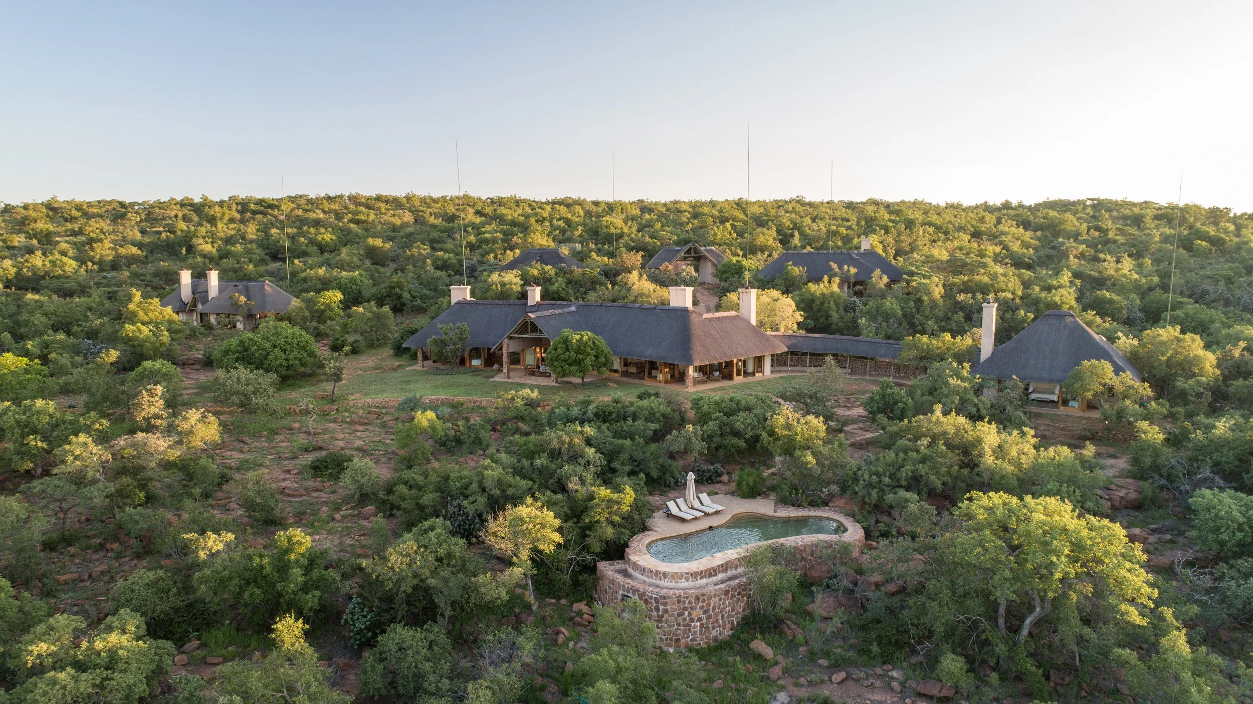 Aerial view of multiple large houses with thatched roofs in a lush, green landscape with trees and shrubs. There is a small swimming pool with poolside lounge chairs in the foreground.