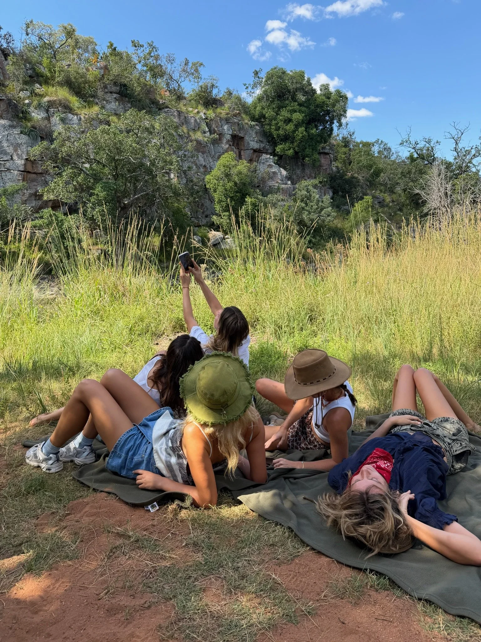 Group of young women lying on blankets outdoors taking photos of the rocky hillside and trees in the background on a sunny day