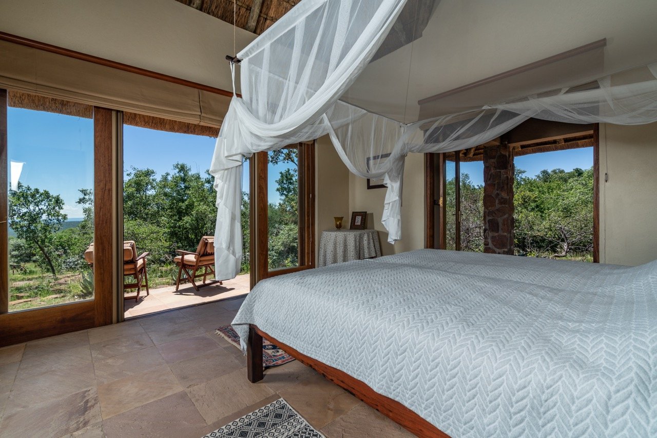 A bedroom with a bed covered in a white quilt, mosquito netting overhead, and a sliding glass door leading to a balcony with outdoor chairs and a view of trees and blue sky.