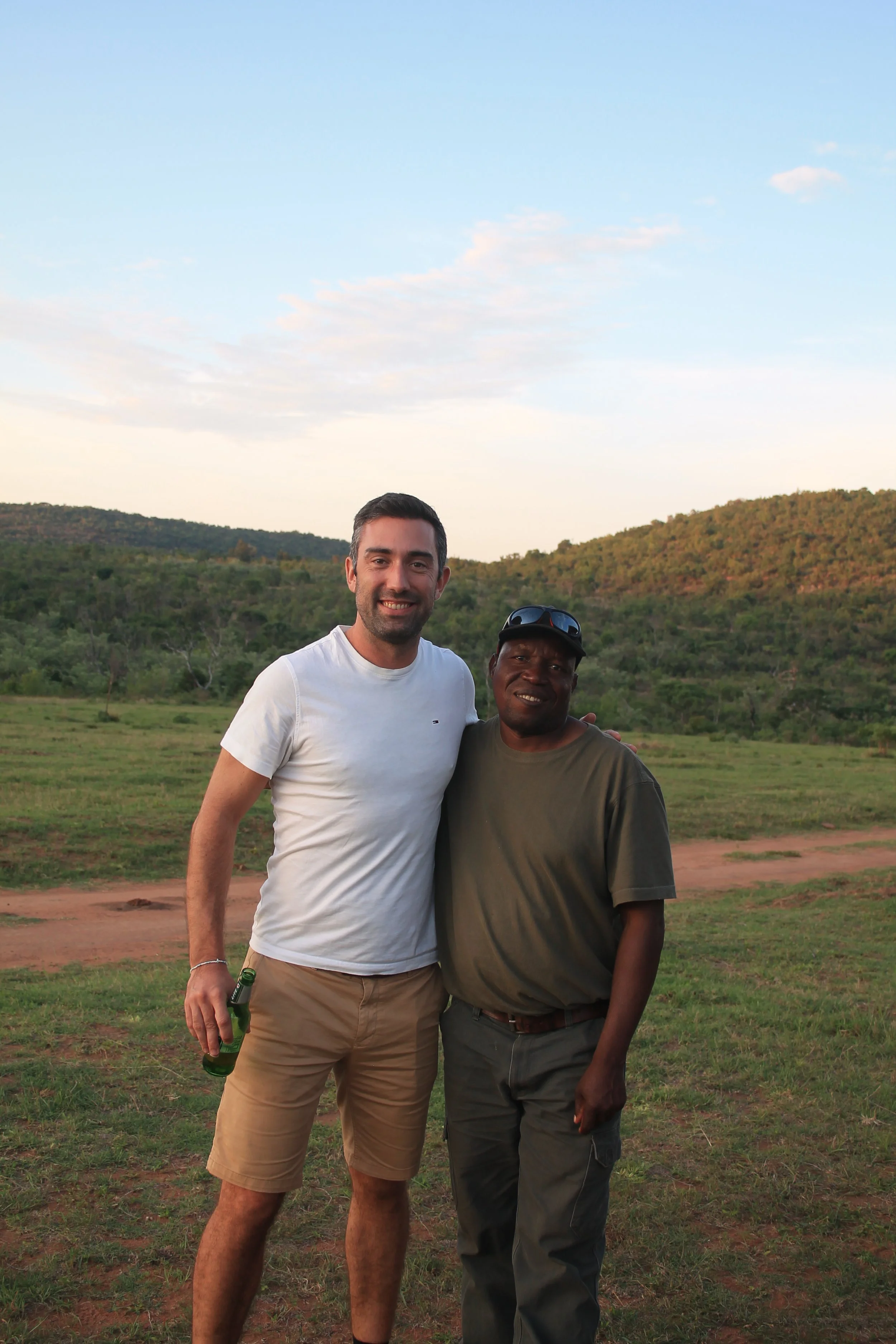 Two men standing outdoors on grass with hills and trees in the background, smiling at the camera.