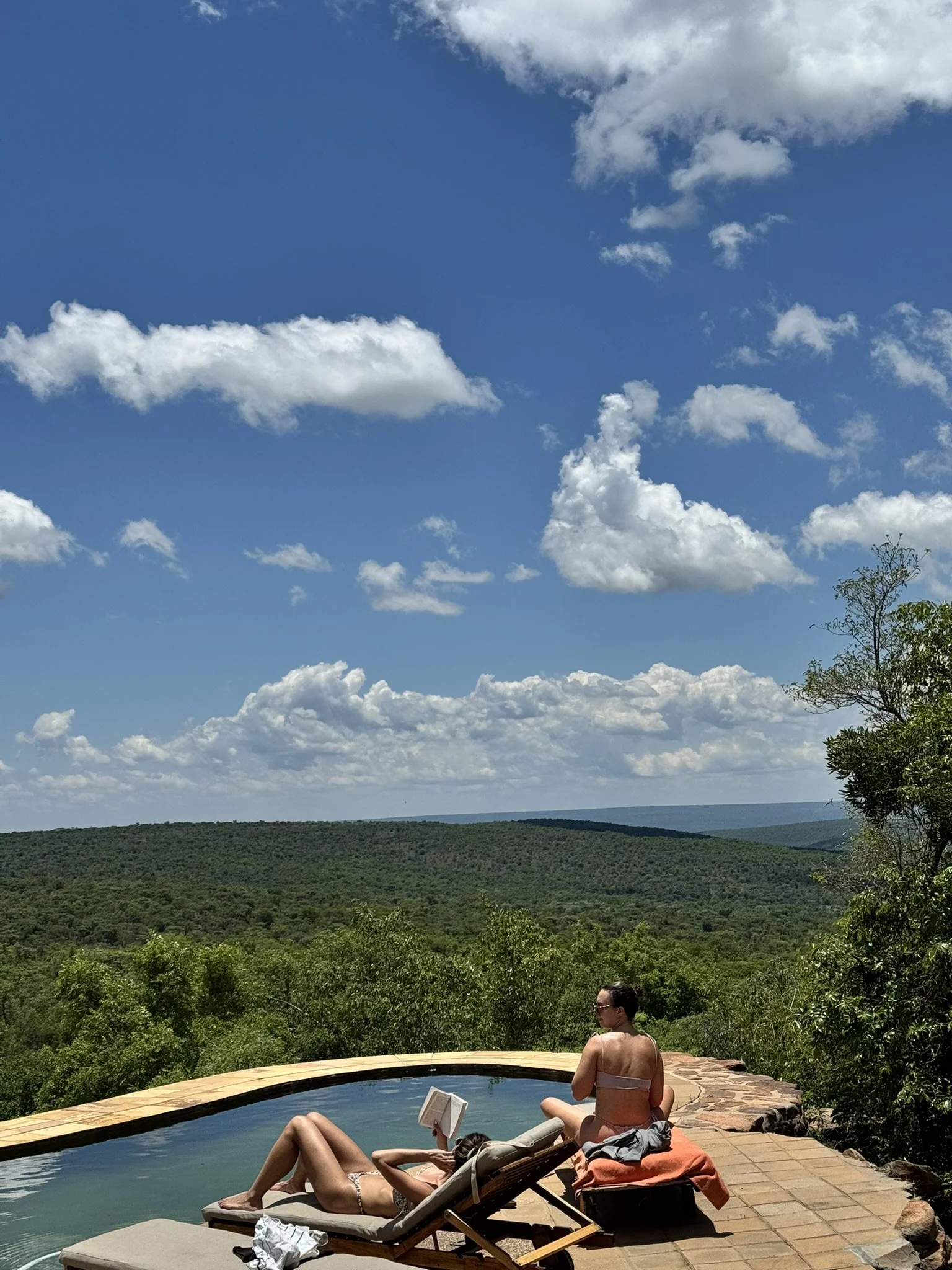 Two women relaxing by a pool with scenic mountain and cloudy sky view, one lying on a lounge chair reading a book and the other sitting on the pool's edge.