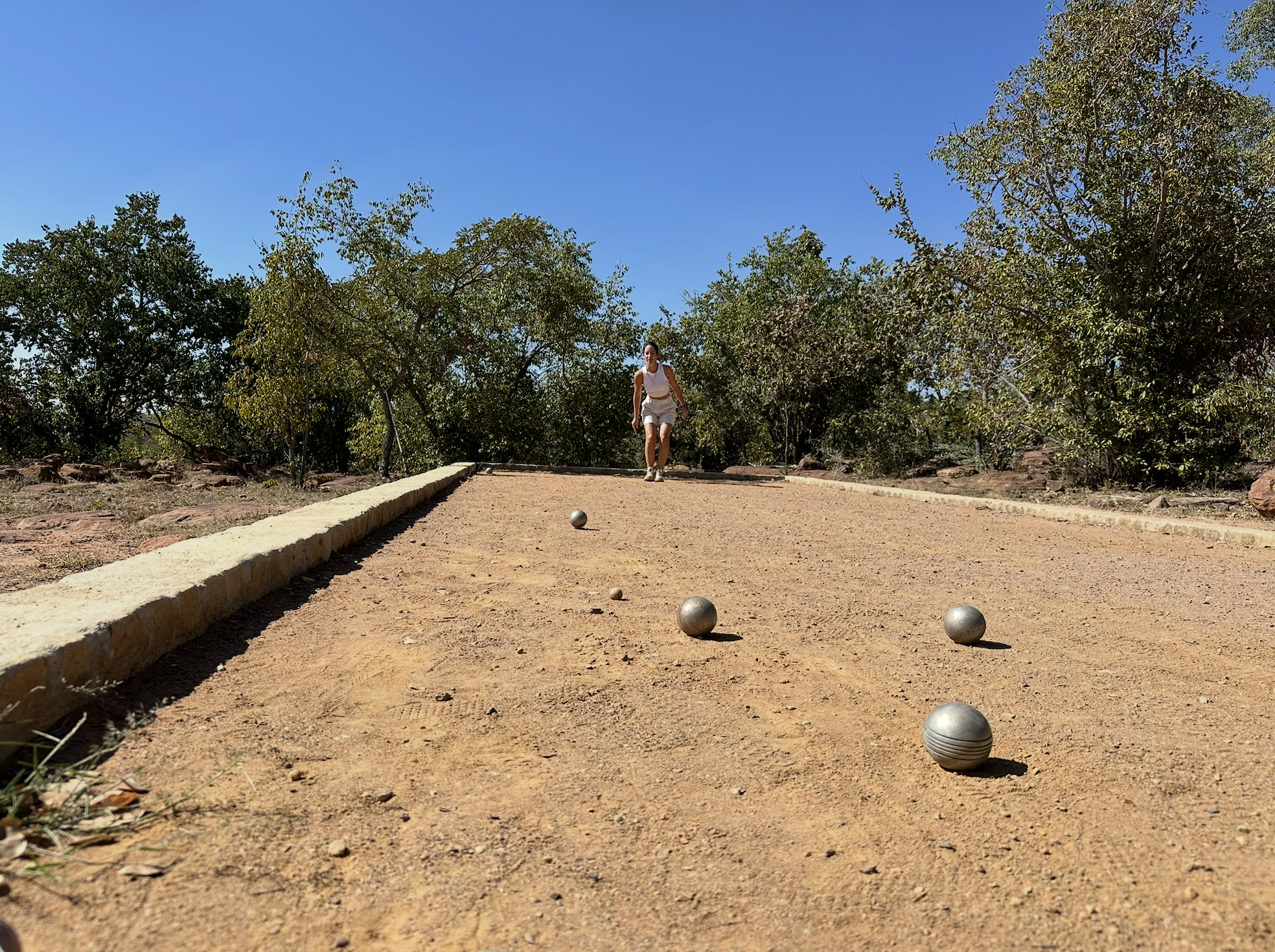 A woman playing lawn bowling on a dirt court outdoors, with trees and a clear blue sky in the background.