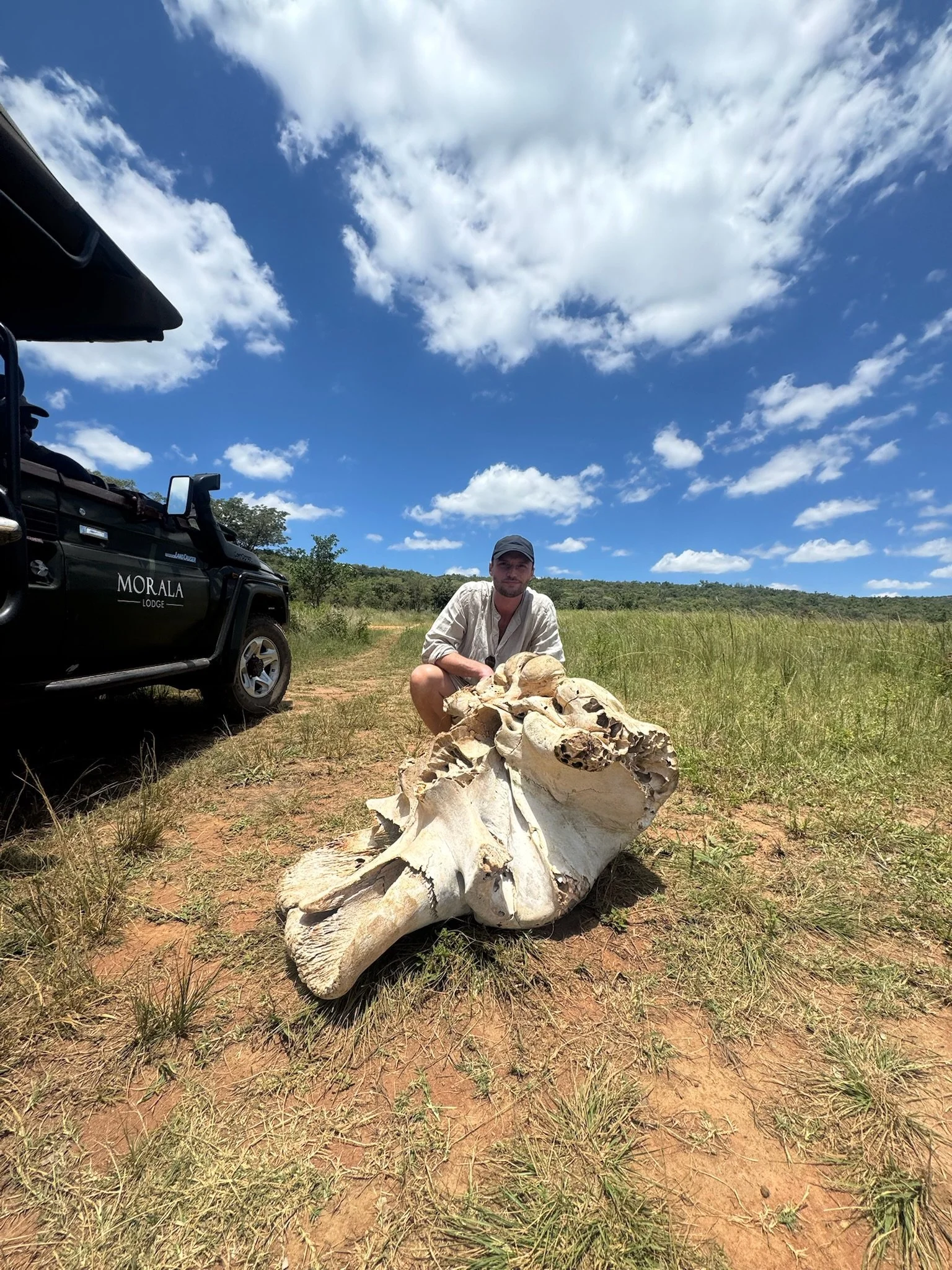 A man squatting next to a large animal skull on a dirt trail in a grassland, with a police-style vehicle labeled 'Morala Lodge' nearby and a blue sky with clouds overhead.