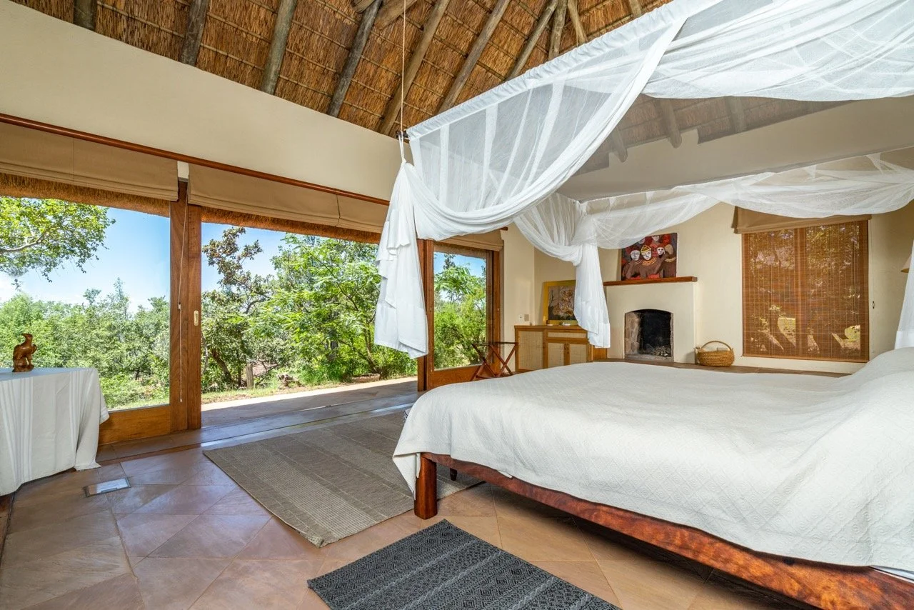 A cozy bedroom with a canopy bed, white bedding, and mosquito netting hanging from a thatched roof. Large glass doors open to a lush green outdoor view. The room features wooden accents, a fireplace, and simple furniture.