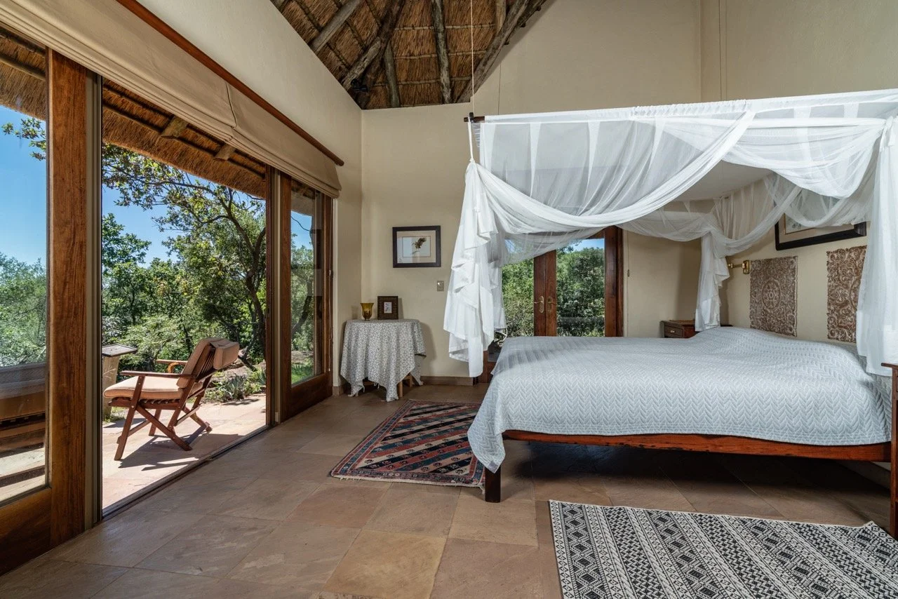 A bedroom with a four-poster bed draped with white fabric, wooden furniture, a sliding glass door leading to an outdoor patio with a chair, and views of green trees.