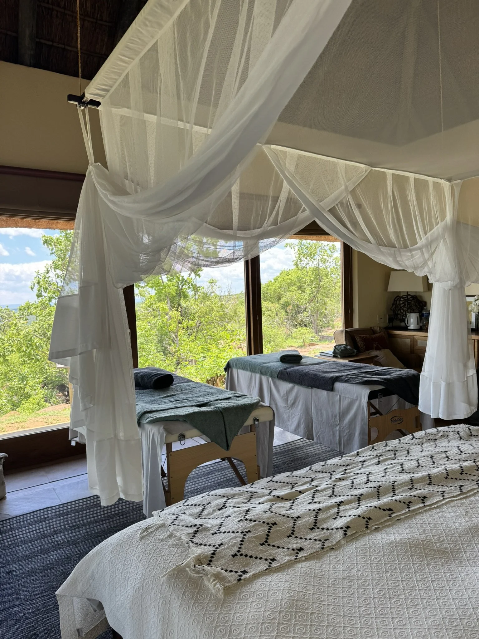 Interior of a spa room with a large bed and two massage tables overlooking a lush green landscape through big windows.