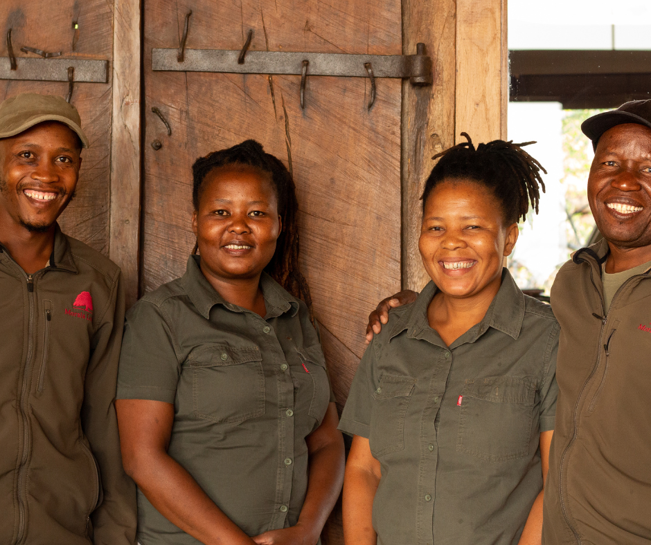 Four people standing indoors, smiling, in front of a wooden wall, wearing green or brown outdoor work jackets.