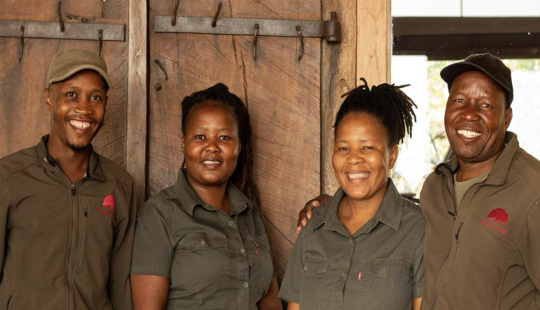 Four smiling people standing together in casual outdoor setting, wearing matching greenish-brown work shirts with red logos, against a wooden background.
