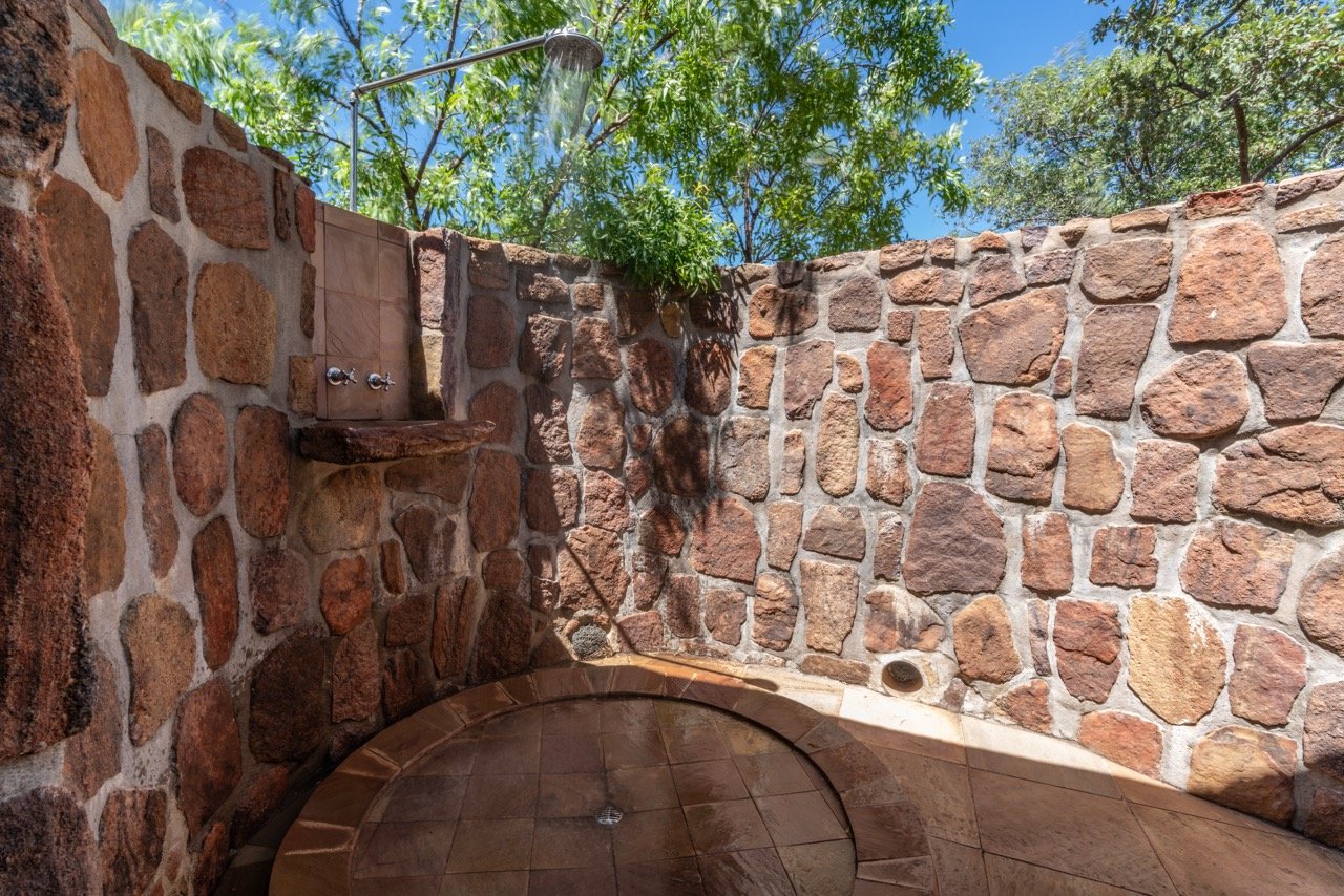 Open outdoor shower with stone walls, a tiled shower floor, and a showerhead attached to the wall. Green trees and blue sky are visible in the background.