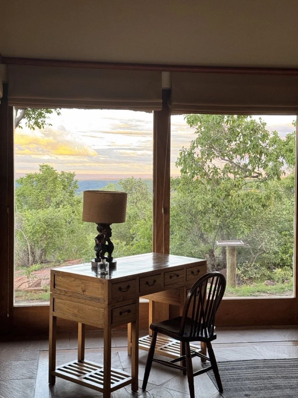 Interior view of a room with a wooden desk and a black wooden chair, a table lamp on the desk, and large windows showing green trees and a sunset sky outside.