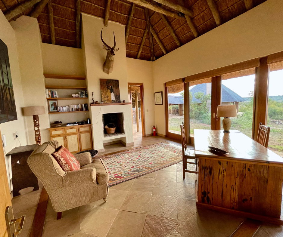 A living room with a thatched ceiling, a fireplace, a large window, and furniture including a sofa, a table, and chairs.