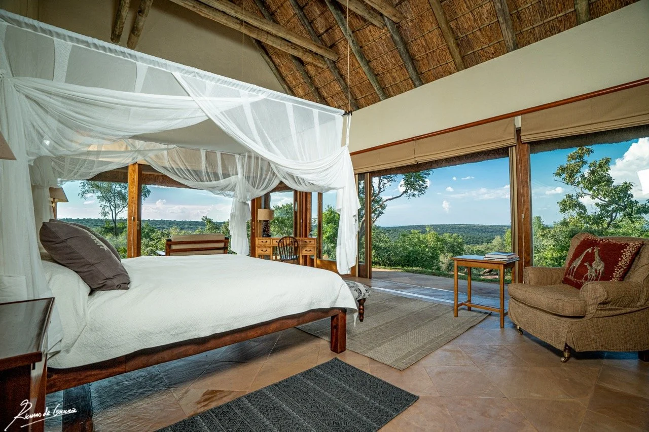 A bedroom with a large bed and a white canopy, large windows with views of trees and a blue sky, a beige armchair with a decorative pillow, a wooden side table with books, and a small writing desk with a chair, all in a rustic style with wooden beams
