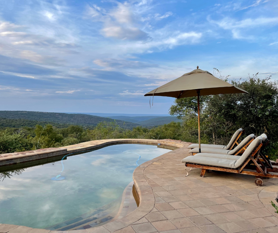 An outdoor swimming pool with a curved edge, two lounge chairs with cushions, and a large beige umbrella. The scene overlooks a landscape of green hills and a partly cloudy sky.