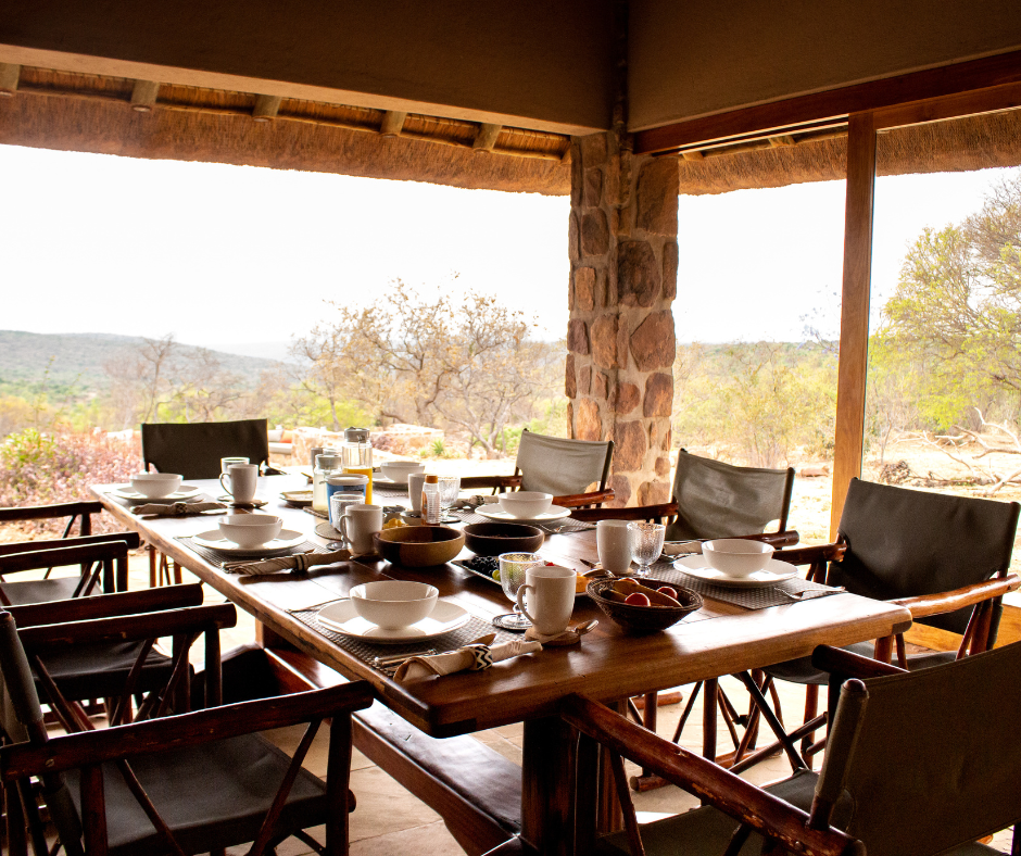 A rustic outdoor dining table set for breakfast with bowls, cups, and plates on a covered patio overlooking a scenic wilderness landscape.