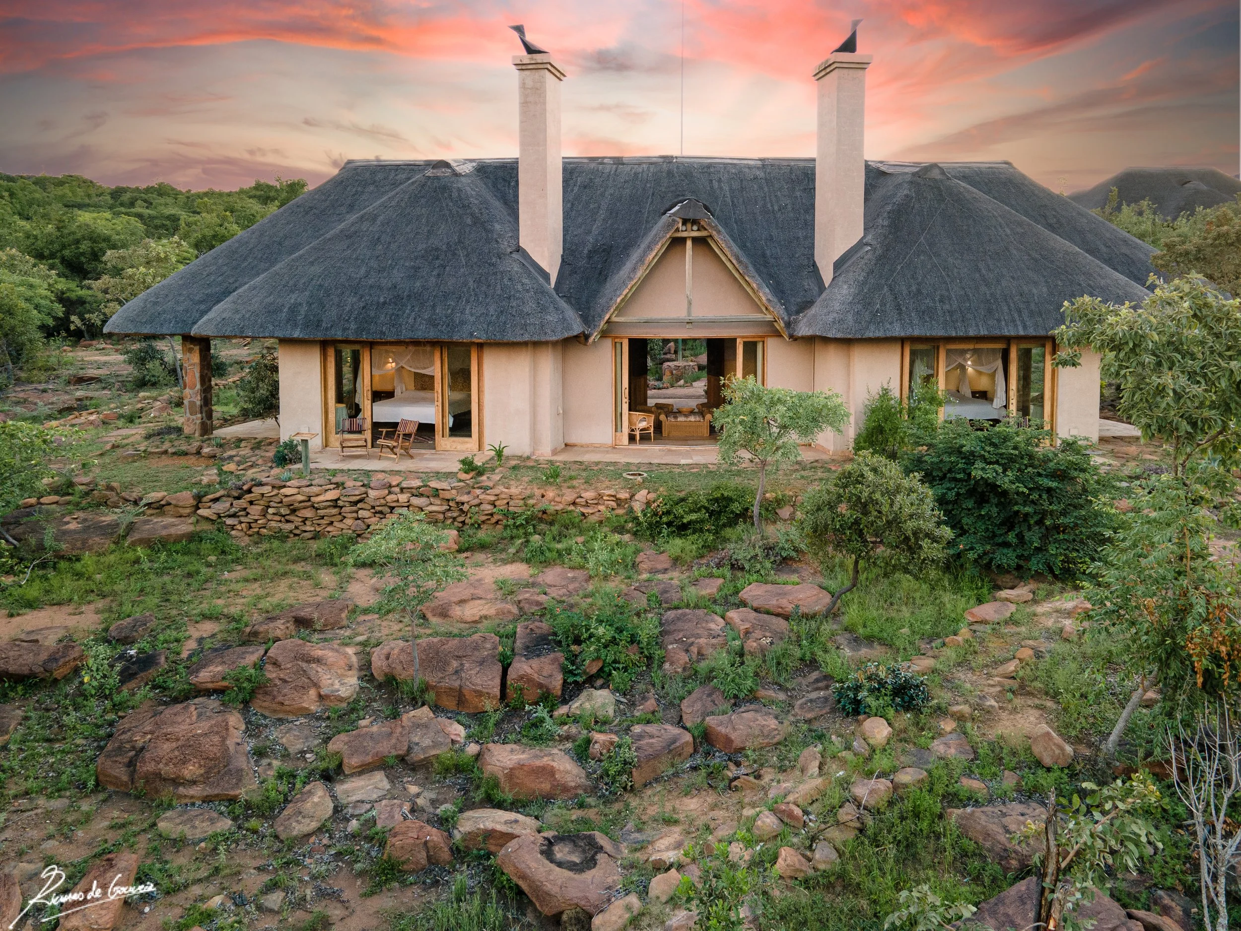A house with a thatched roof and large glass windows on a rocky and green landscape at sunset.