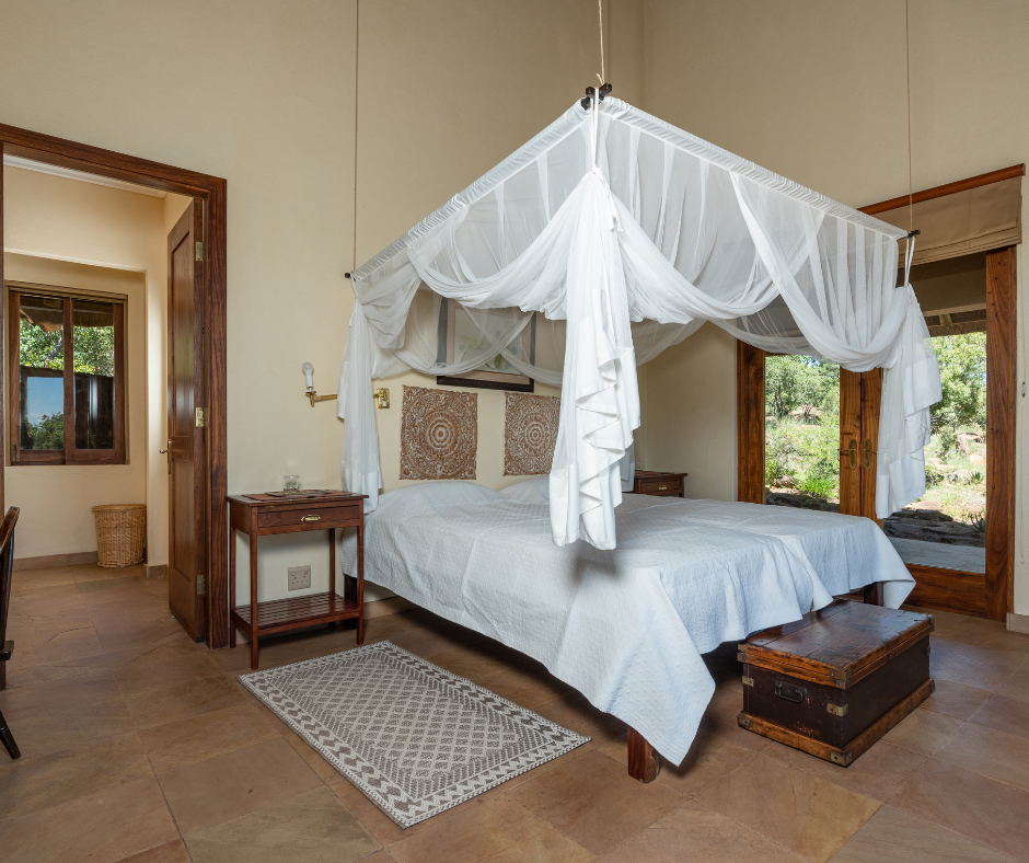 A bedroom with a four-poster white canopy bed, wooden side tables, and large windows showing greenery outside.