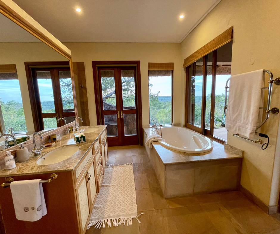 Bathroom with double vanity sinks, a large mirror, a tile countertop, and a bathtub near large windows with a view of trees and a cloudy sky outside.