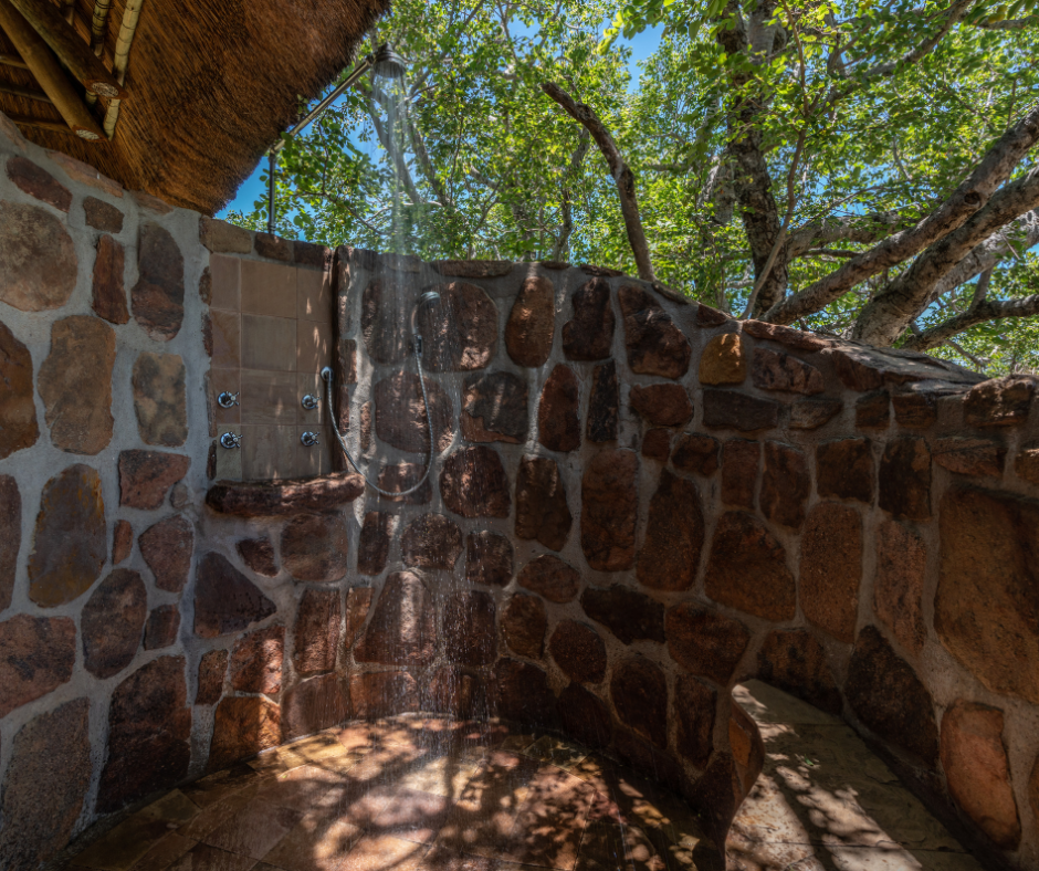 Outdoor stone shower with a roof, surrounded by trees and greenery, with sunlight filtering through.