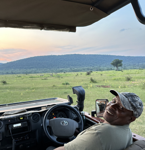 A smiling man wearing a camo hat sits in the driver’s seat of a vehicle, with a vast green field and distant hills in the background, possibly on a safari or outdoor excursion.