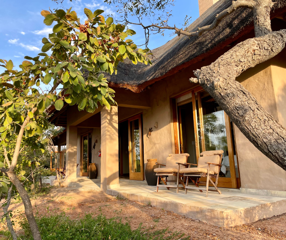 A house with a thatched roof, large glass doors, and a small porch with two chairs. A large tree leans over the porch, partially shading it during sunset.