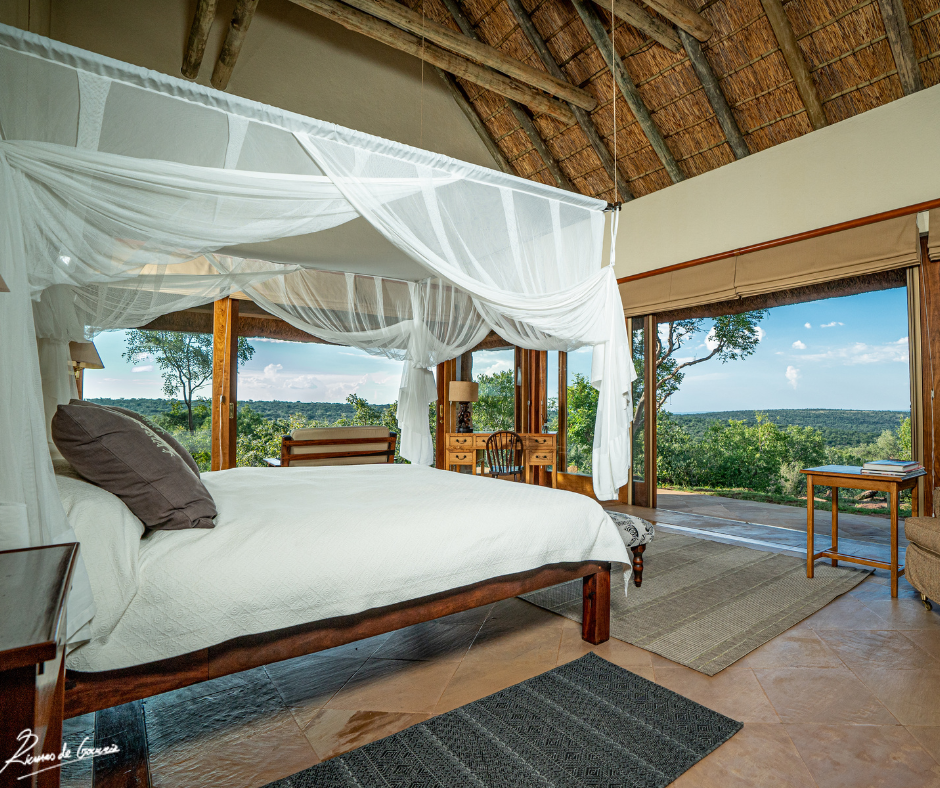 A cozy bedroom with a wooden canopy bed and white drapes, overlooking a lush green landscape through large glass windows.