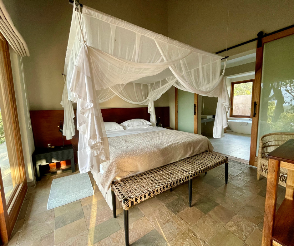 A bedroom with a bed draped in a white canopy, a dark wood headboard, white bedding, a woven bench at the foot of the bed, a small rug, and windows showing trees outside. There is also a view into a bathroom with a freestanding bathtub and sink.