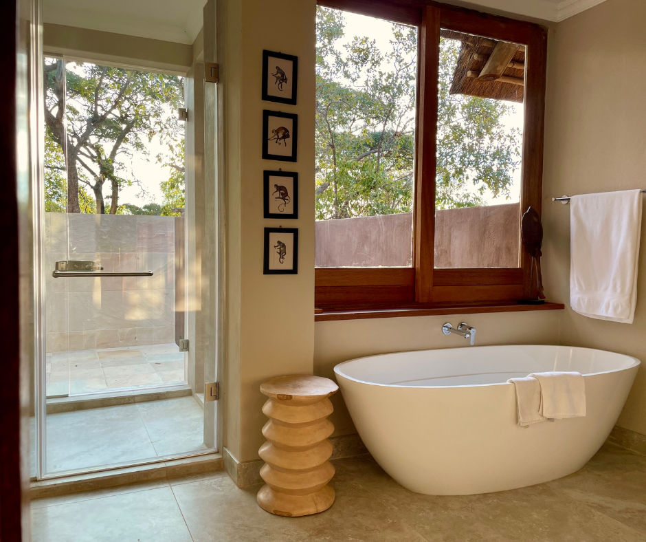A bathroom with a white bathtub next to a large window showing trees outside, a glass shower door to the left, a towel on a towel rack on the right, and a small wooden stool beside the tub.