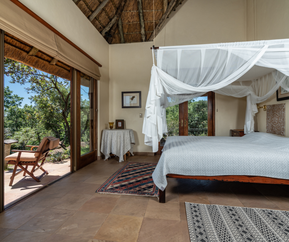 A bedroom with a large bed, white bedding, and a canopy. There is a sliding glass door leading to an outdoor patio with a chair, and views of trees outside. The room has a thatched roof and wooden furniture.