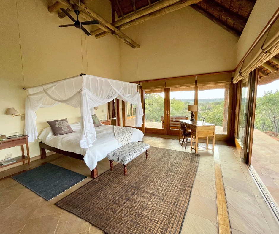 A spacious bedroom with a large four-poster bed draped with white fabric, wooden side tables, a bench at the foot of the bed, a desk and chair near large sliding glass doors, and a view of trees and open sky outside.