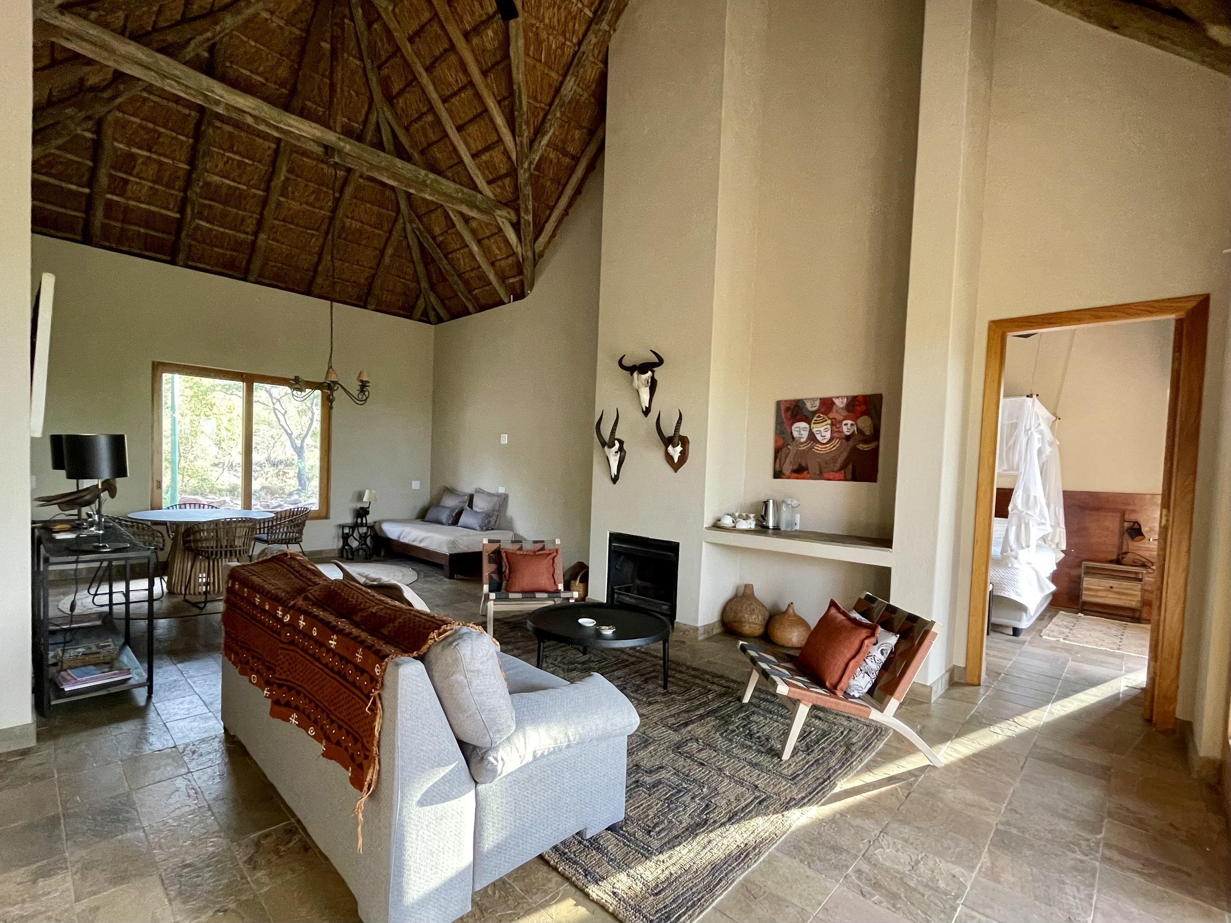 Living room with a high vaulted wooden ceiling, beige walls, and tiled floor. Furniture includes a light gray sofa with a colorful throw, a patterned lounge chair with pillows, a small round black table, and multiple chairs and a table near the window. Decor includes mounted animal skulls above the fireplace, wall art, and decorative vases. A doorway shows a bedroom with a white bedspread.
