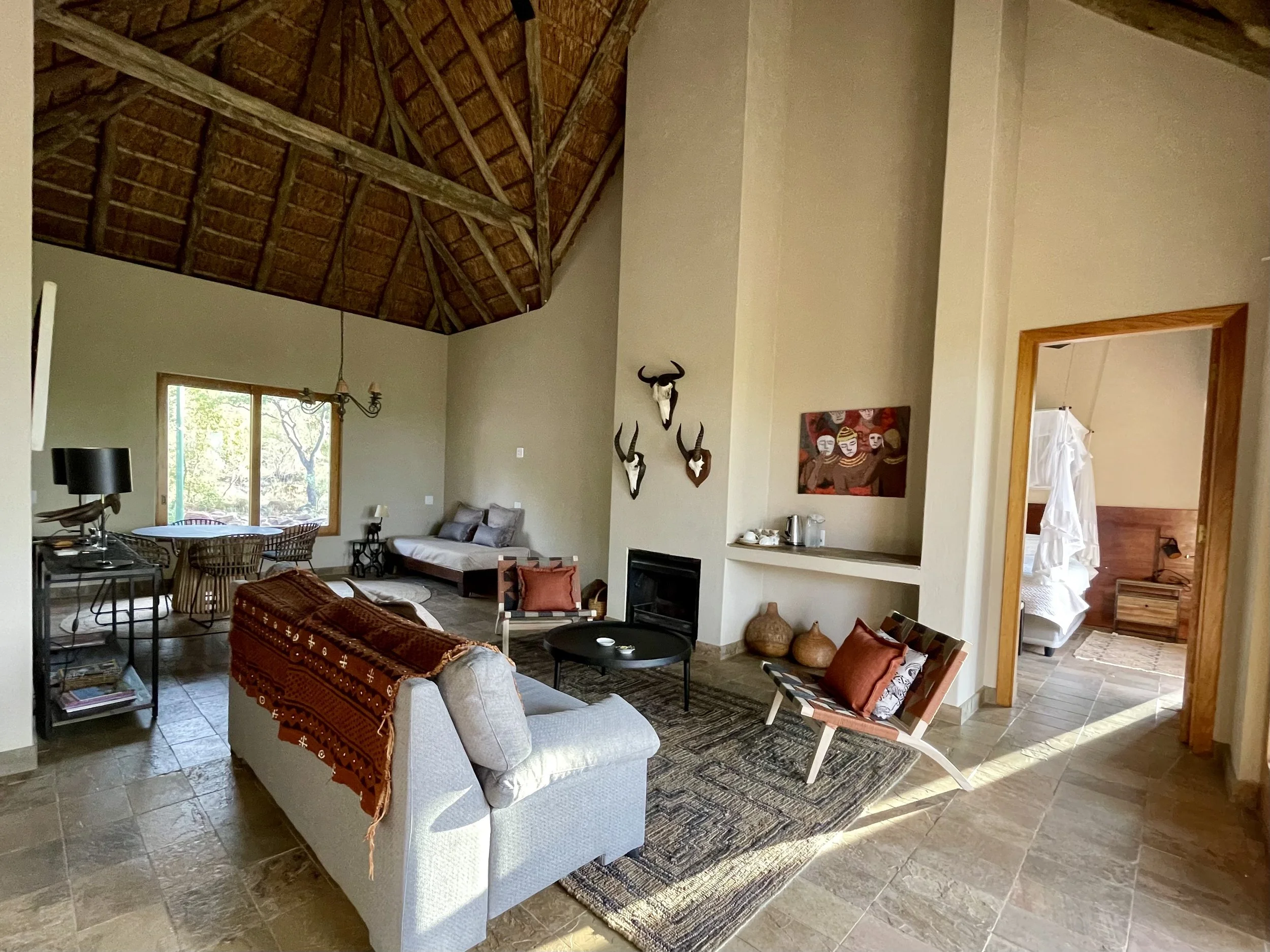 Living room with high vaulted wooden ceiling, beige walls, and tiled floor. Contains a light gray sofa with a red throw, a black round coffee table, a colorful armchair with pillows, and a fireplace on the wall. Decor includes mounted animal skulls a