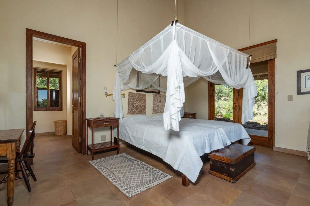 A bedroom with a four-poster bed with white drapes, wooden furniture, a small rug, and windows showing greenery outside.