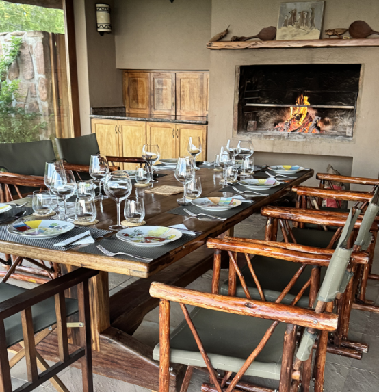 Dining room with a long wooden table set for dinner, featuring plates, wine glasses, and utensils, surrounded by wooden chairs, with a fireplace in the background and a kitchen area with wooden cabinets.