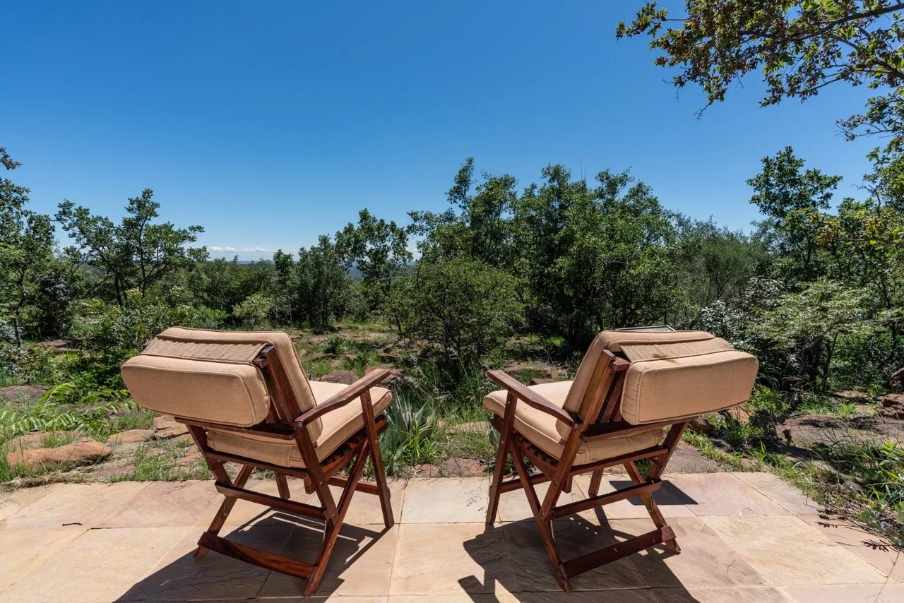 Two beige cushioned patio chairs with wooden frames face a scenic view of trees and greenery under a clear blue sky.